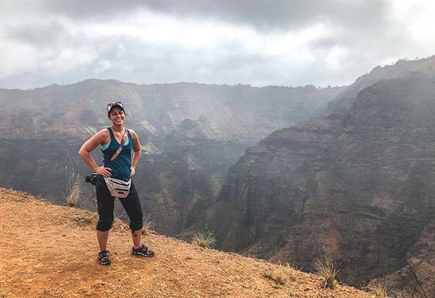 Brittany smiling close to the ledge of an overlook with beautiful mountains in the background on a hike to Waipo'o Falls where they were engaged in Kauai Hawaii